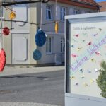 Po prawej stronie gablota z napisem radosnej wielkanocy, kolorowymi papierowymi ptaszkami i bukszpanem. W tle żółta kamienica. Na pierwszym planie rozmazane kolorowe jajka robione na szydełku