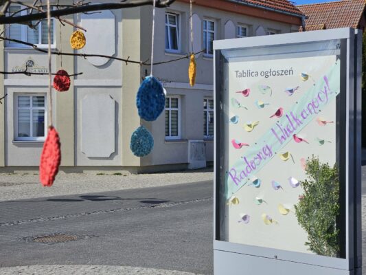 Po prawej stronie gablota z napisem radosnej wielkanocy, kolorowymi papierowymi ptaszkami i bukszpanem. W tle żółta kamienica. Na pierwszym planie rozmazane kolorowe jajka robione na szydełku